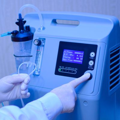 Hands in medical gloves hold out an oxygen mask against the background of the oxygen generator concentrator to deliver at low lung oxygen saturation for Covid-19 pneumonia. Focus on oxygen mask